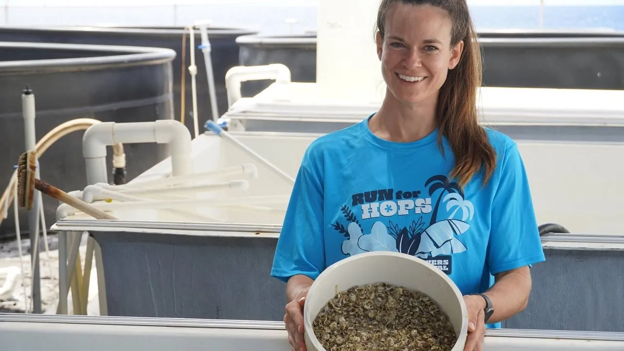Woman holding a bucket of oysters beside aquaculture tanks.