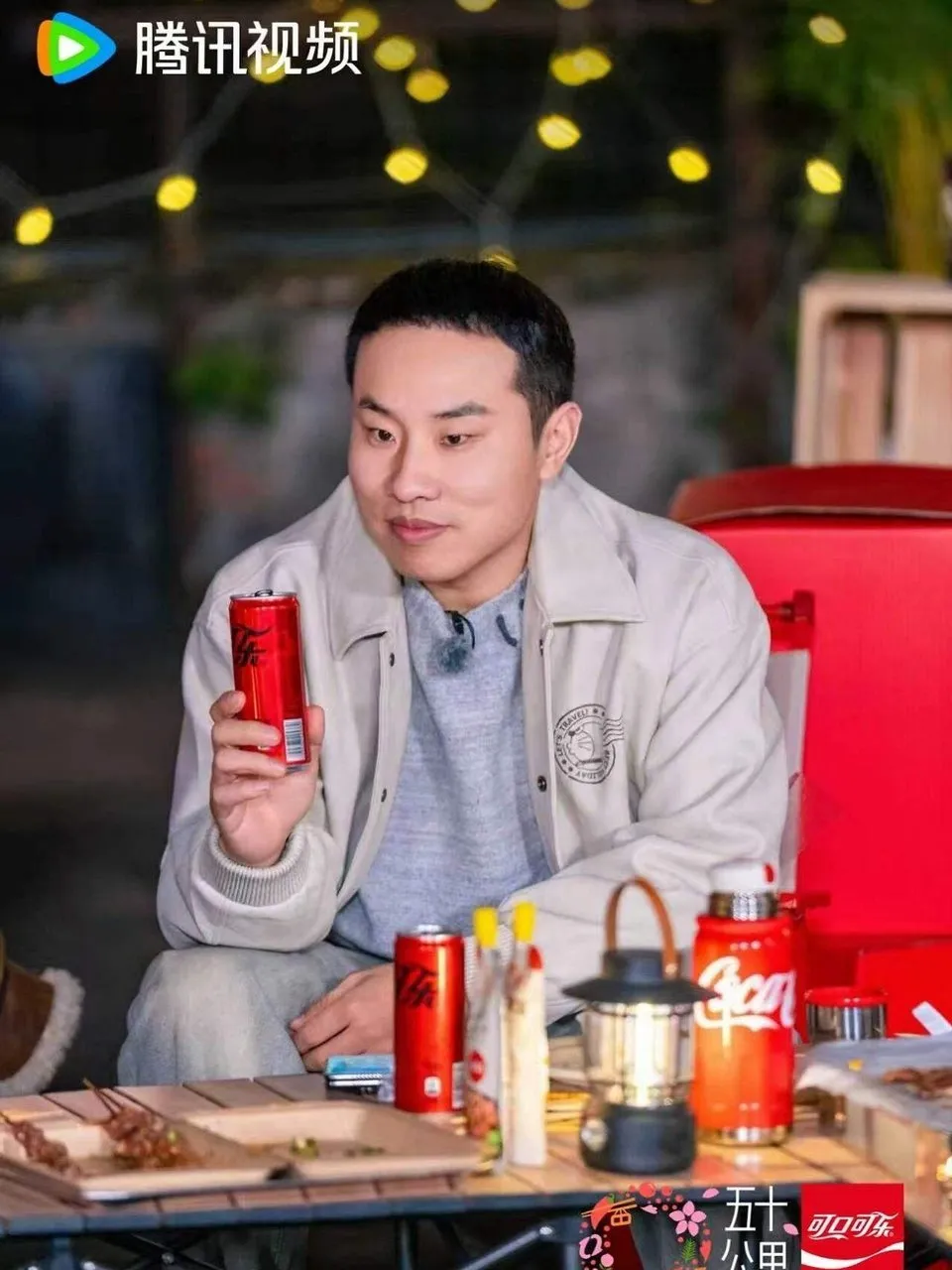 Man holding a red soda can at an outdoor table with food, lantern, and string lights.