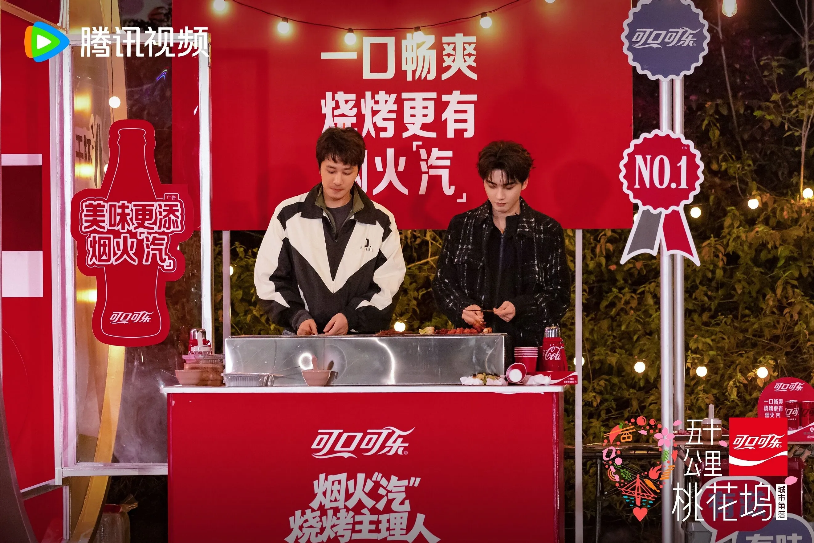 Two men grill skewers at a red Coca-Cola promotional stall.