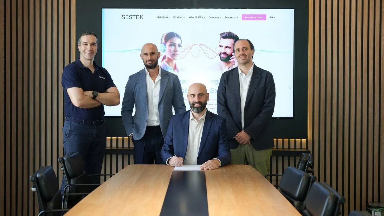 Four men in a boardroom, with one seated signing a document in front of a SESTEK screen.