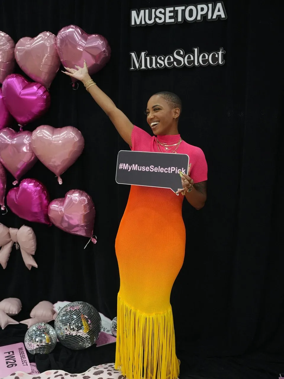 Woman in a pink and orange fringed dress holding a #MyMuseSelectPick sign beside heart balloons.