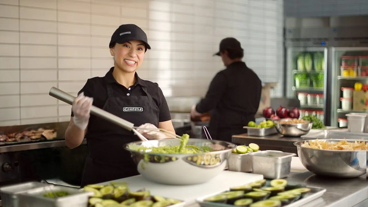 Chipotle employee serving guacamole beside bowls of avocado, limes and tortilla chips in a kitchen.