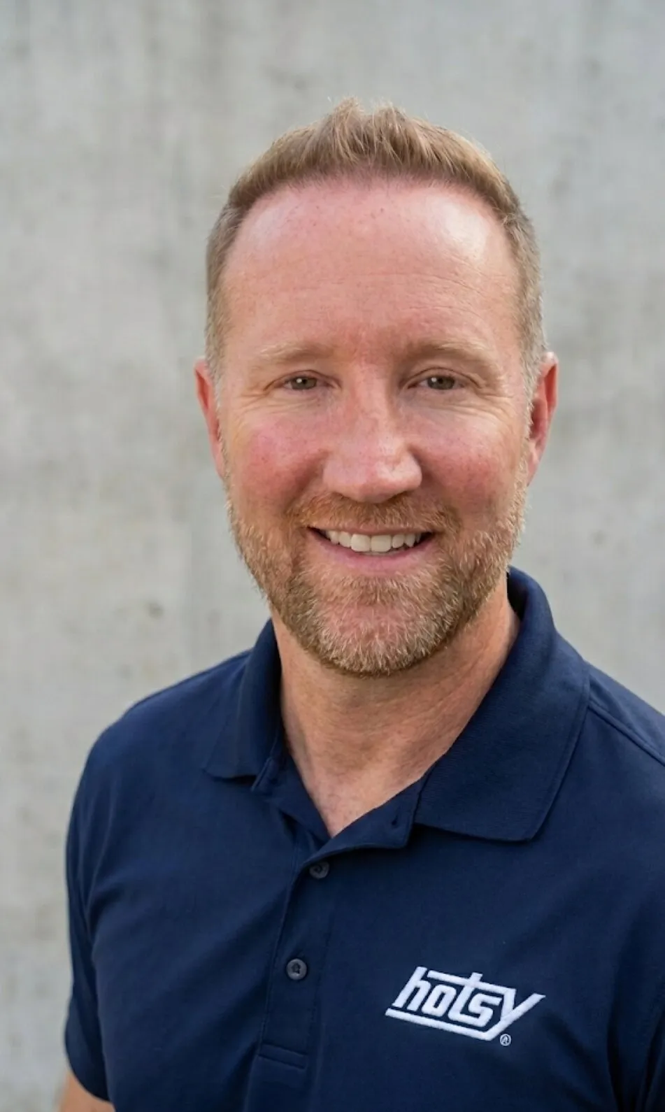 Aaron Muller smiling in a navy polo shirt against a gray wall.