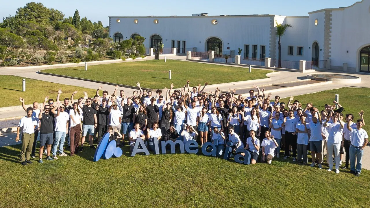 A large group of employees waving beside an Almedia sign on a lawn outside a white building