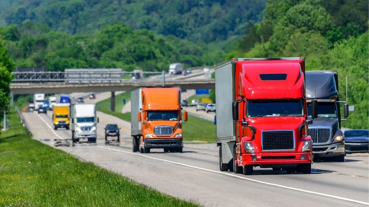 Tractor-trailers and cars traveling on a tree-lined highway beneath an overpass.