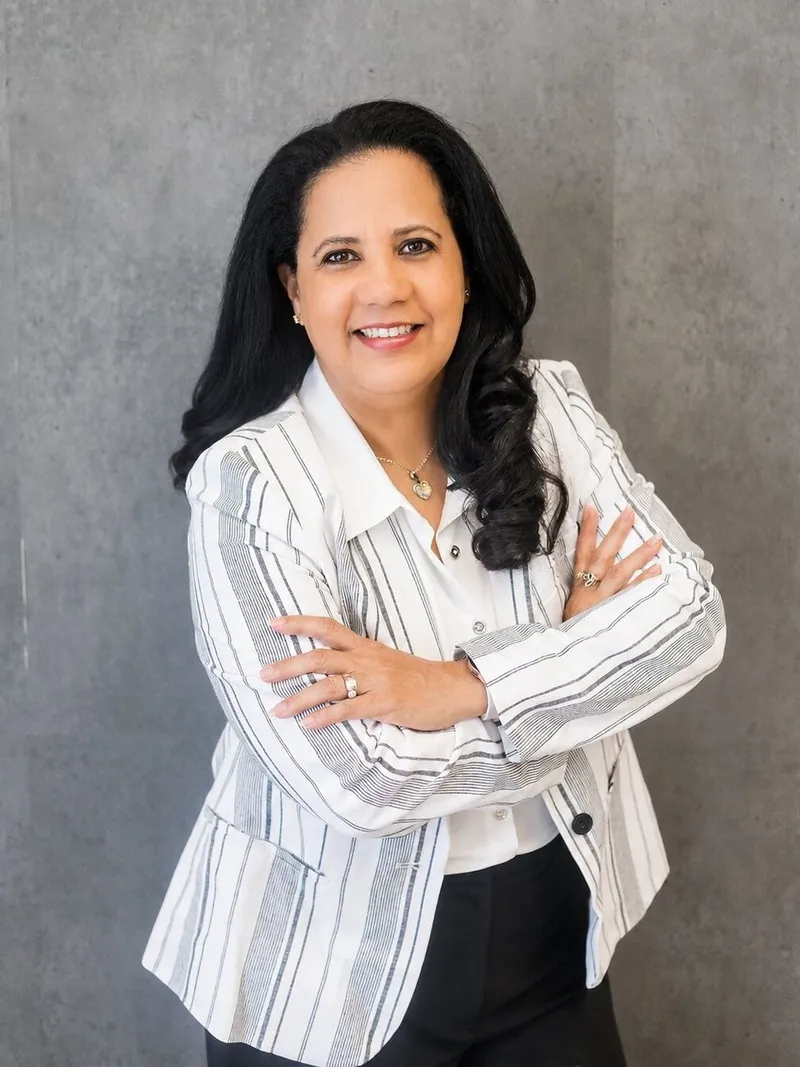 Aicha Bascaro in a white striped blazer, smiling with arms crossed against a gray backdrop.