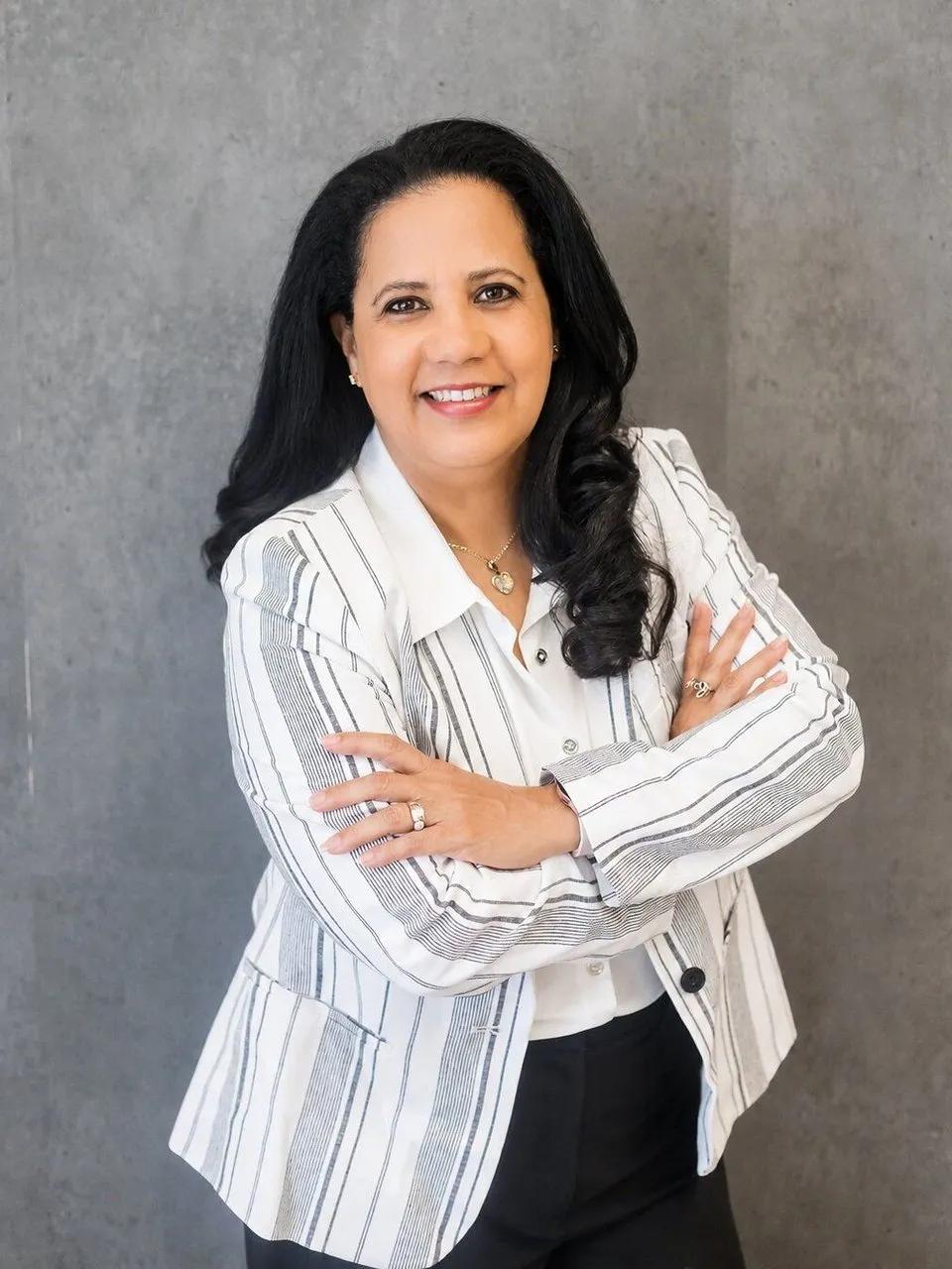 Aicha Bascaro in a white striped blazer, smiling with arms crossed against a gray backdrop.