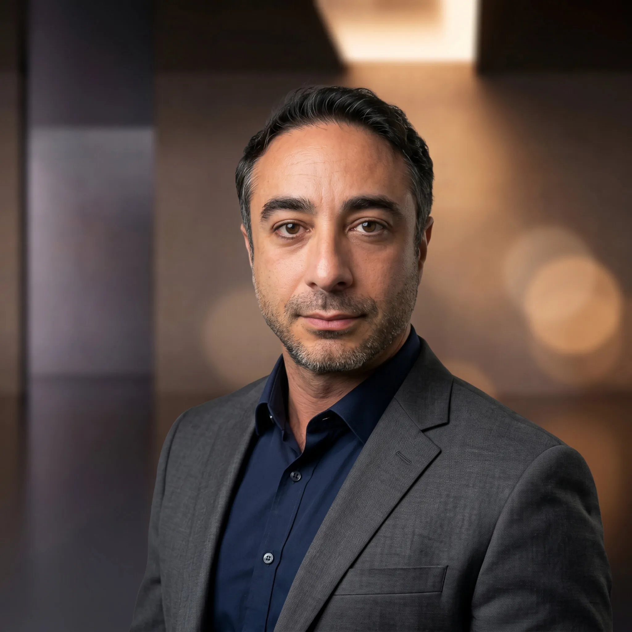 A man in a gray suit jacket and dark blue shirt against a blurred indoor background.
