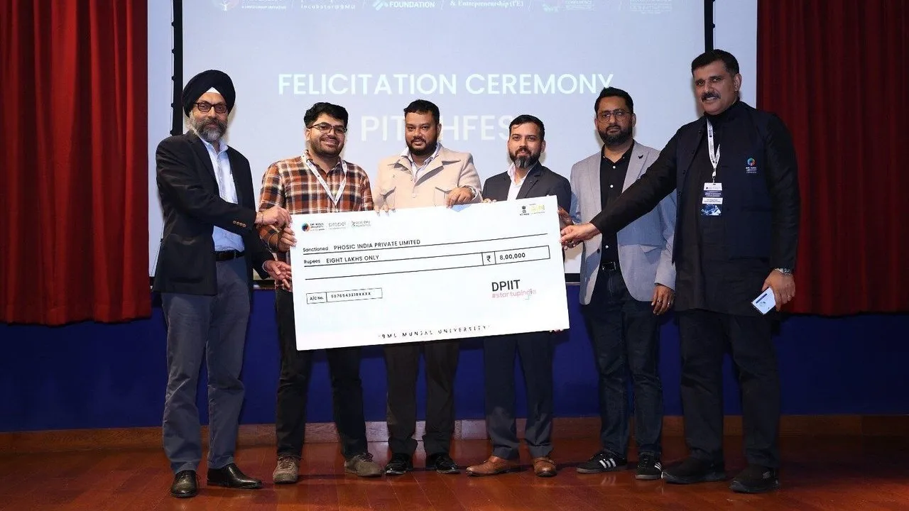 Six men hold a ceremonial cheque for ₹8 lakh on a stage.