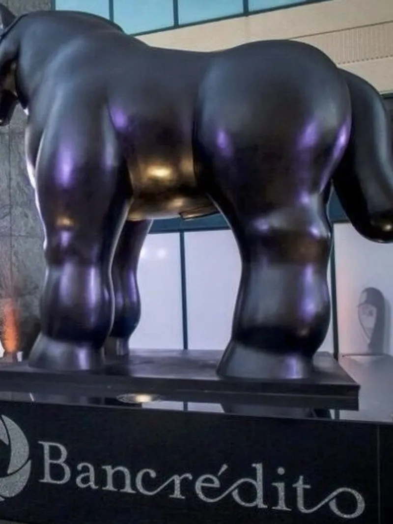 A black, rounded horse sculpture on a Bancroédito pedestal outside a building.