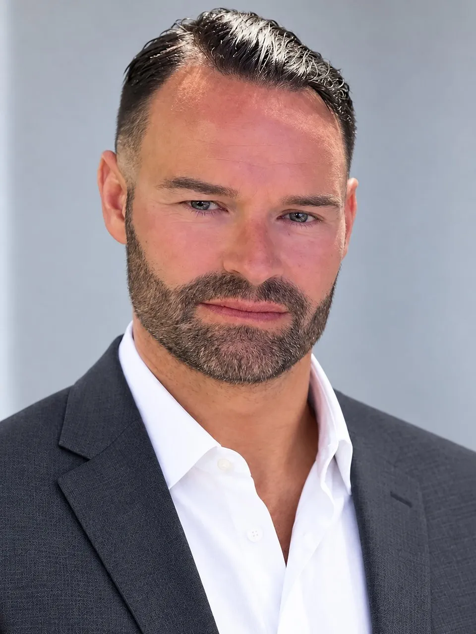 A bearded man in a dark suit jacket and white shirt against a gray background.