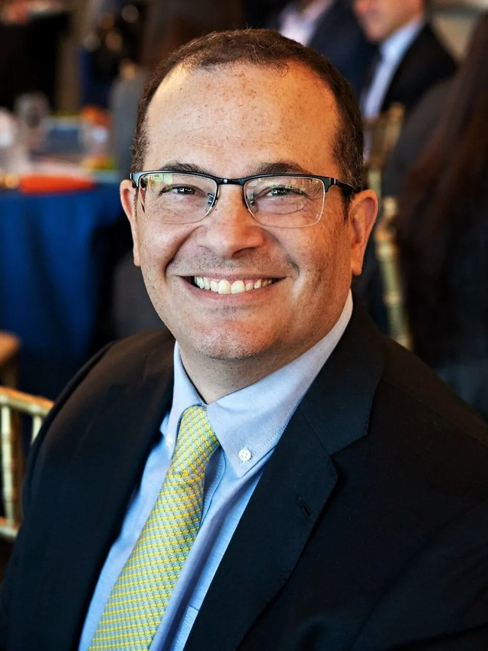 Carl Mazzanti in a dark suit, blue shirt and yellow tie, smiling at an event.