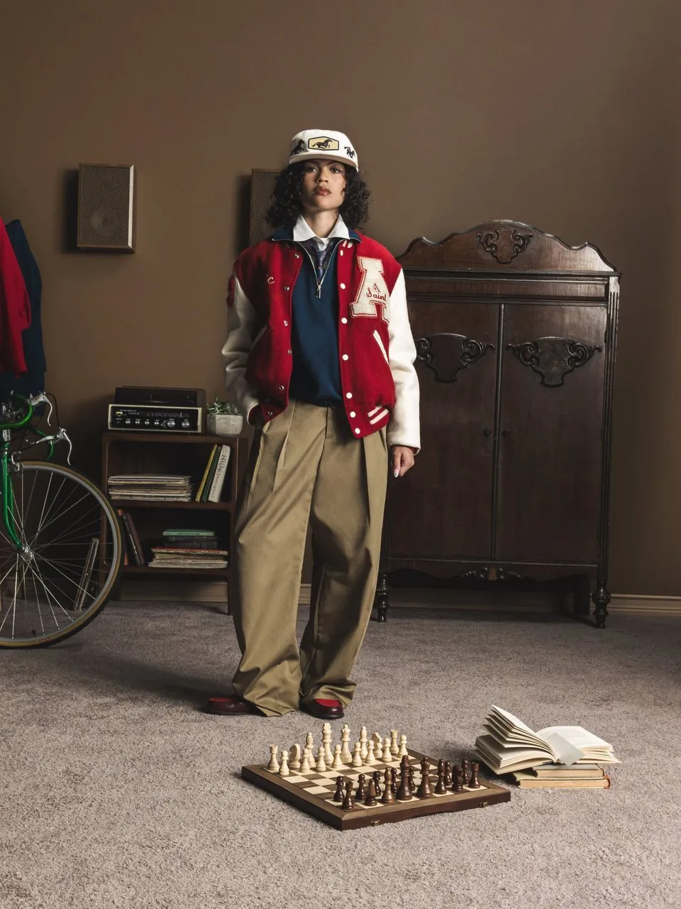 Person in varsity jacket and cap stands beside a wardrobe, bicycle, chessboard and books.