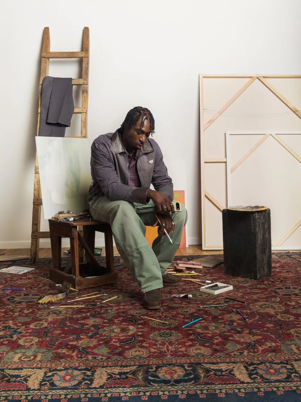 A man in workwear sits in an art studio surrounded by canvases, brushes and pencils.