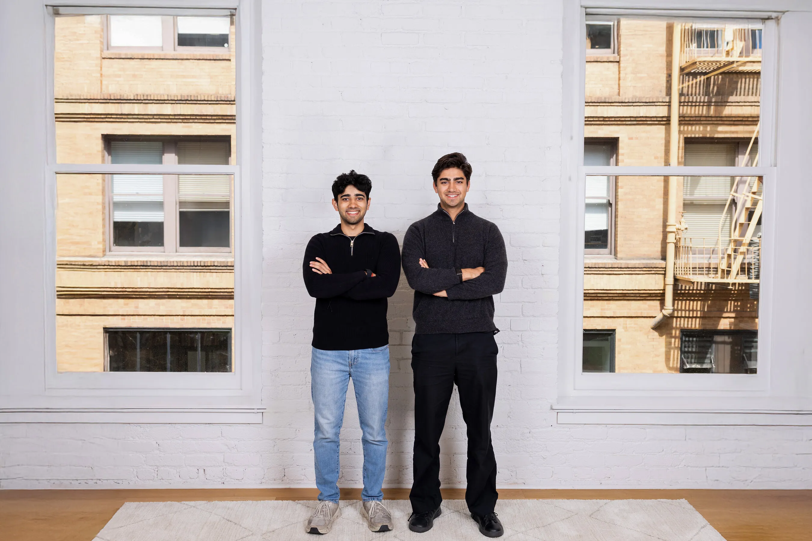 Two men standing with folded arms against a white brick wall between large windows.