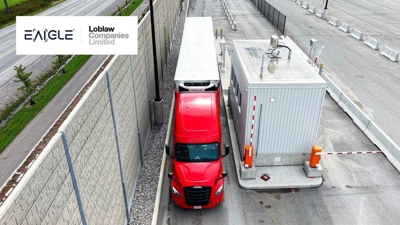 Red semi-truck passing a gatehouse at a secured facility entrance.