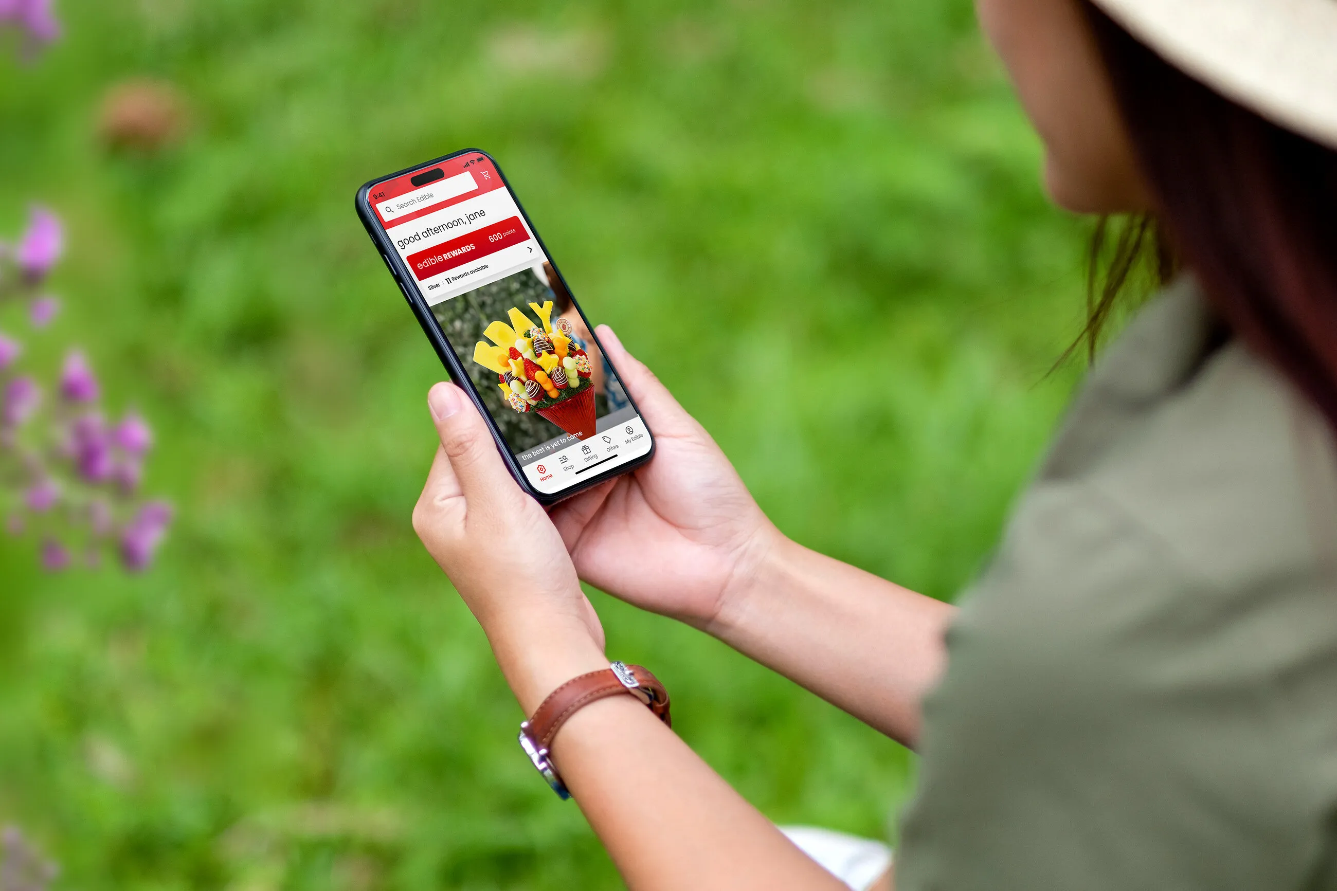 Person holding a phone displaying the Edible Arrangements app and a fruit bouquet against a grassy background