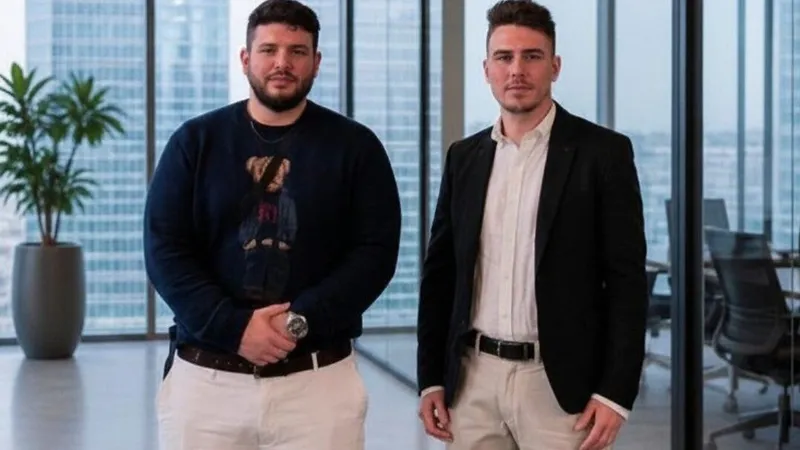 Two men standing in an office with floor-to-ceiling windows and a potted plant.