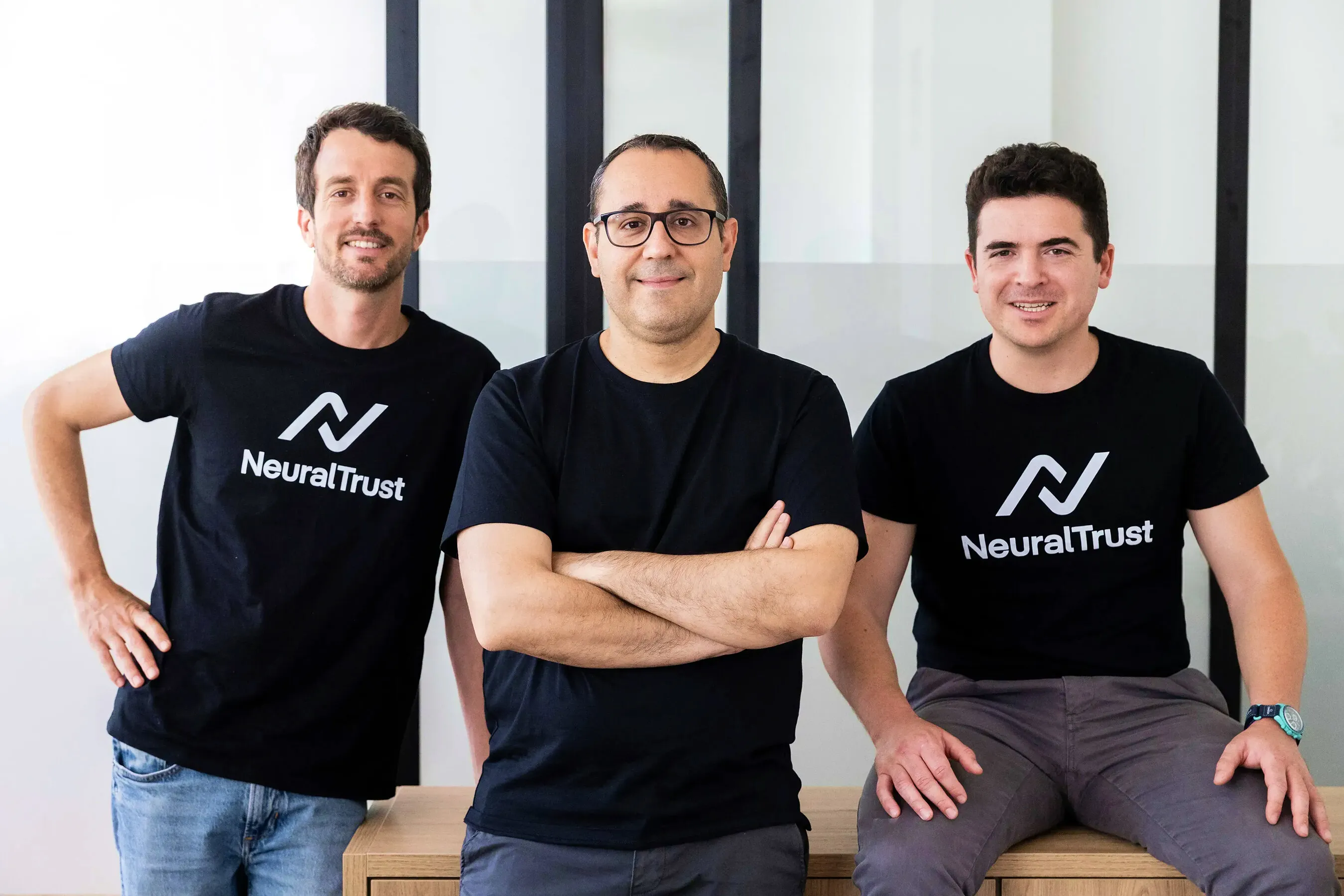 Three NeuralTrust founders wearing black shirts, standing and sitting in an office.