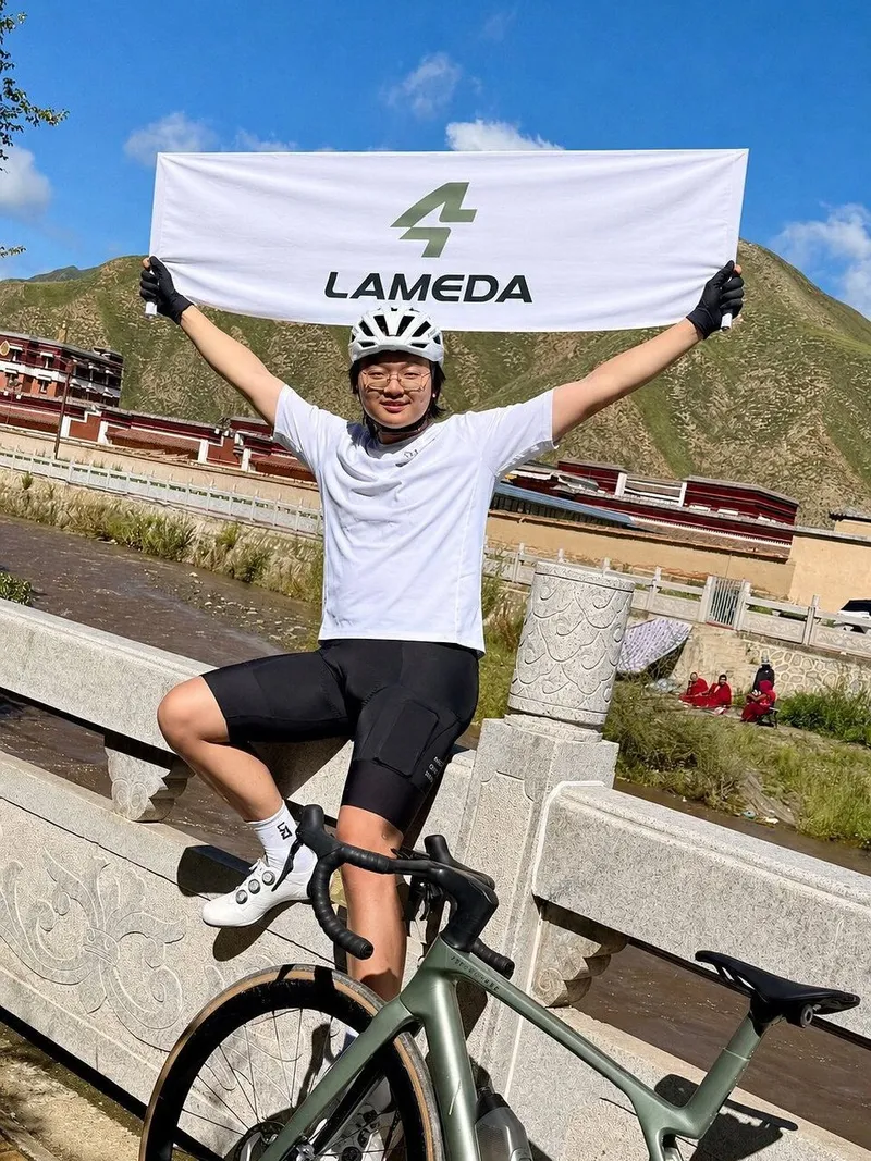 Cyclist holding a LAMEDA banner above a road bike beside a river, with mountains and buildings behind.