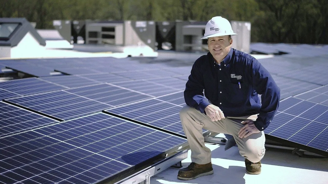 A worker in a hard hat kneels beside rooftop solar panels.