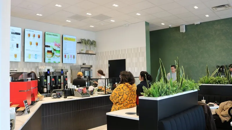 Customers at a coffee shop counter with menu screens, espresso machines and indoor plant dividers.