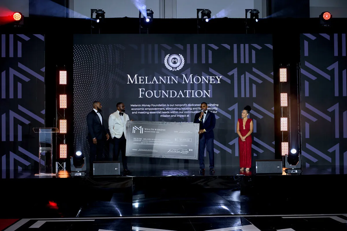 Four people onstage present a large ceremonial check before a Melanin Money Foundation backdrop.
