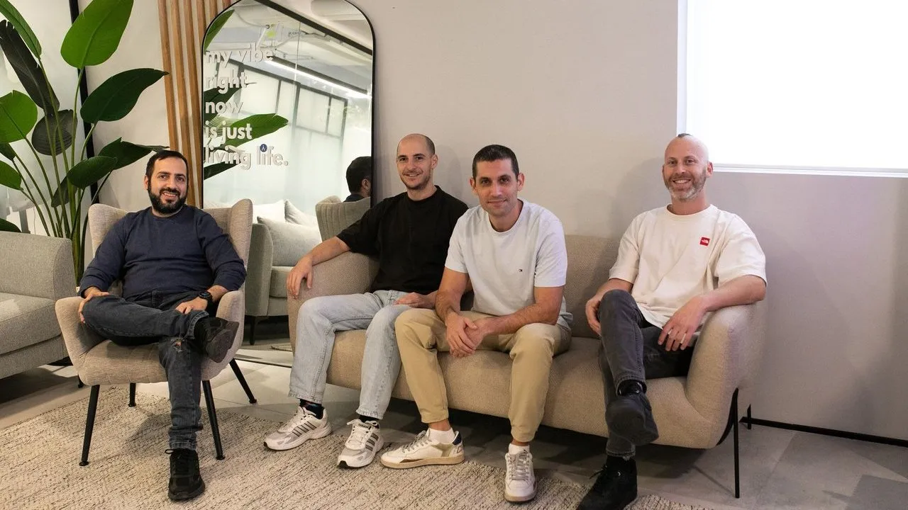 Four men seated on armchairs and a sofa in a modern office lounge beside a large plant and mirror.