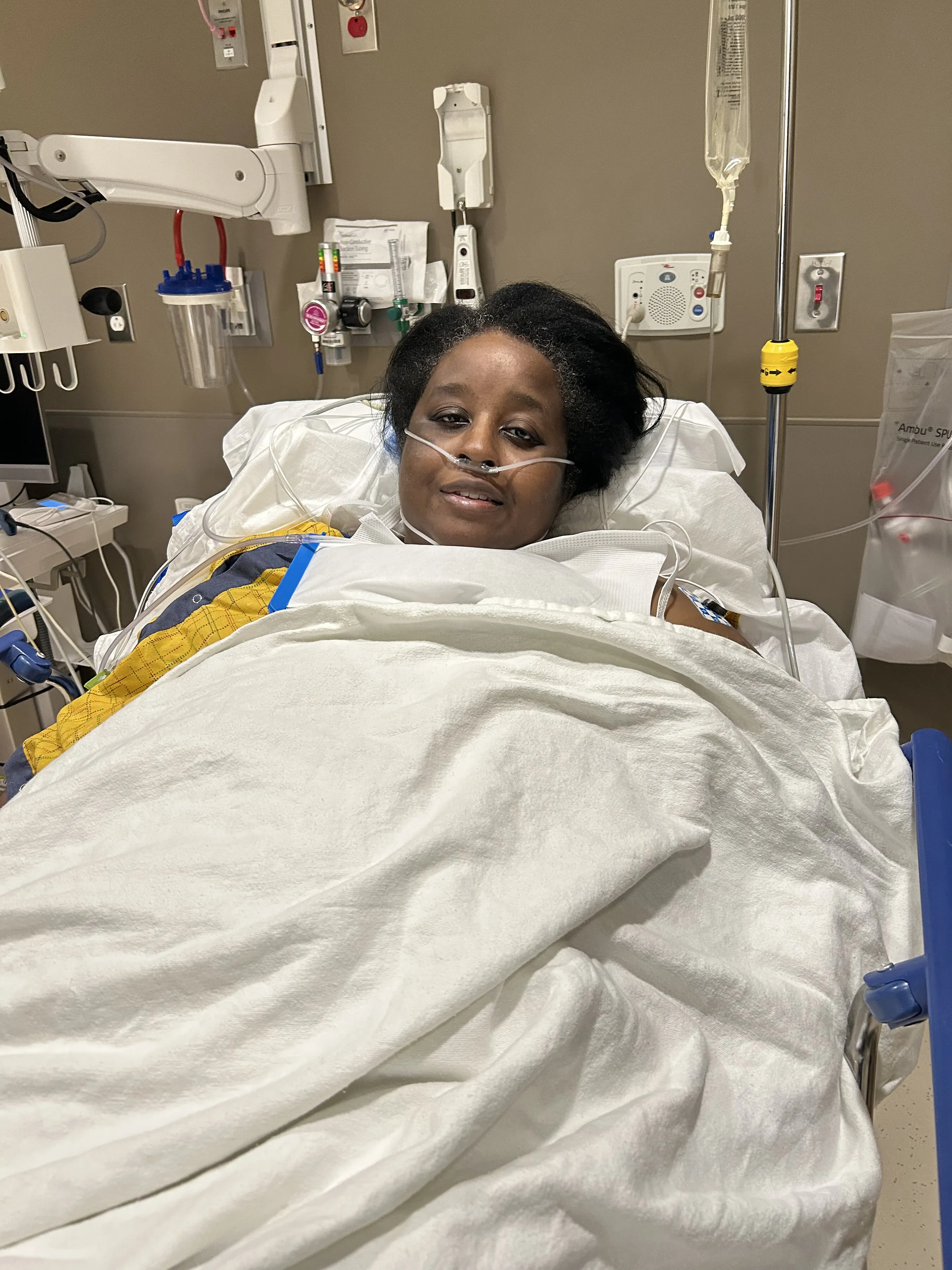 A woman lies in a hospital bed with a nasal oxygen tube and medical equipment nearby.