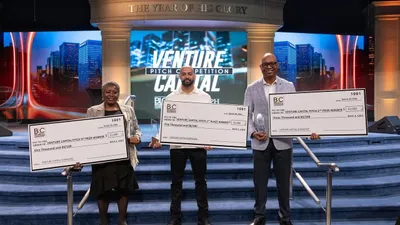 Three winners hold oversized prize checks and trophies on a stage.