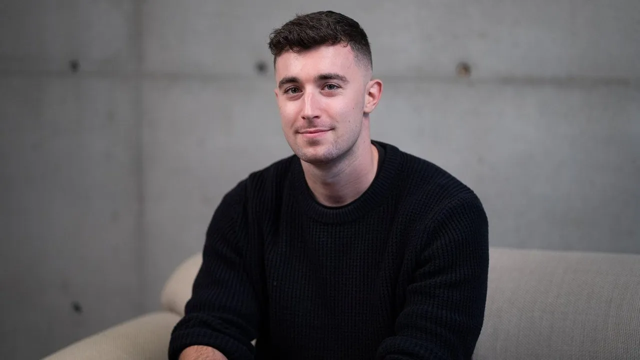 A man in a black sweater sits in a beige chair against a gray concrete wall.