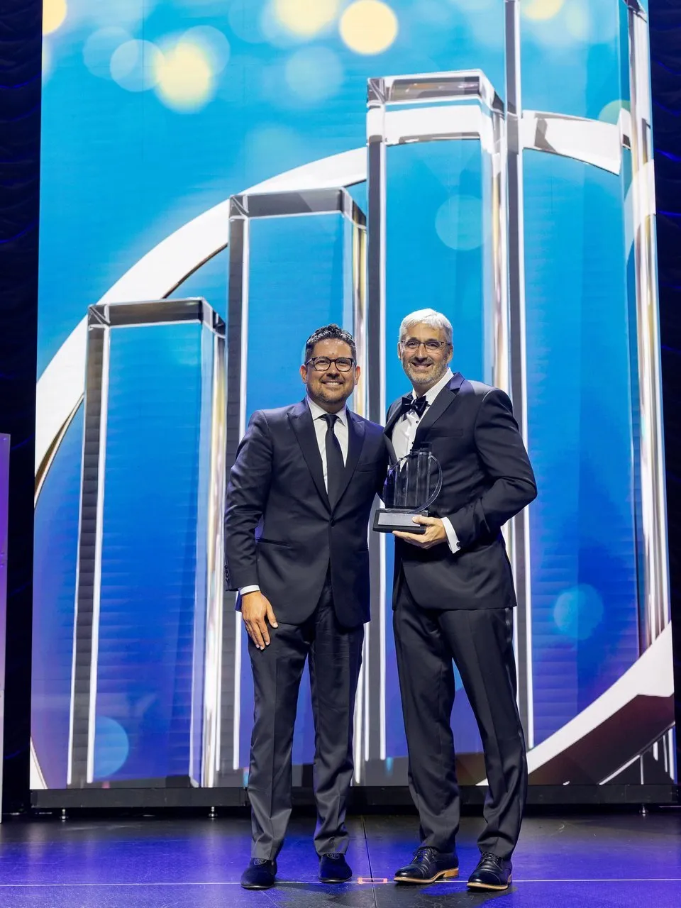 Two men in formal suits onstage, one holding a glass award.