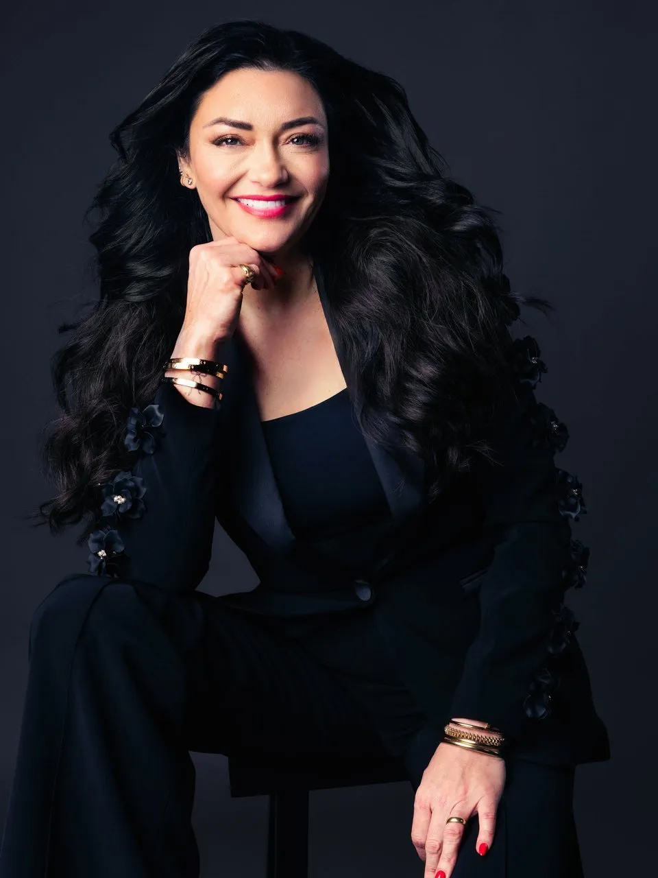 Smiling woman with long dark hair in a black suit, seated against a dark background.