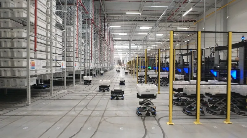 Autonomous robots move bins through a warehouse with tall storage racks and fenced machinery.
