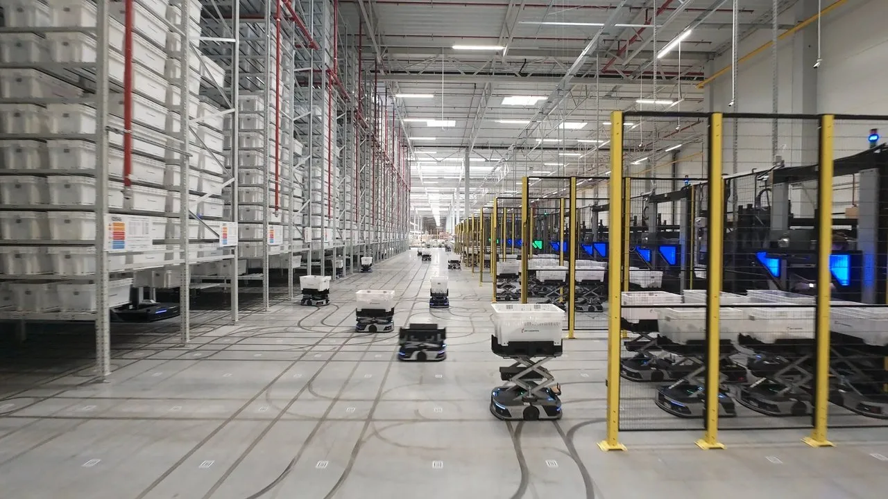 Autonomous robots move bins through a warehouse with tall storage racks and fenced machinery.