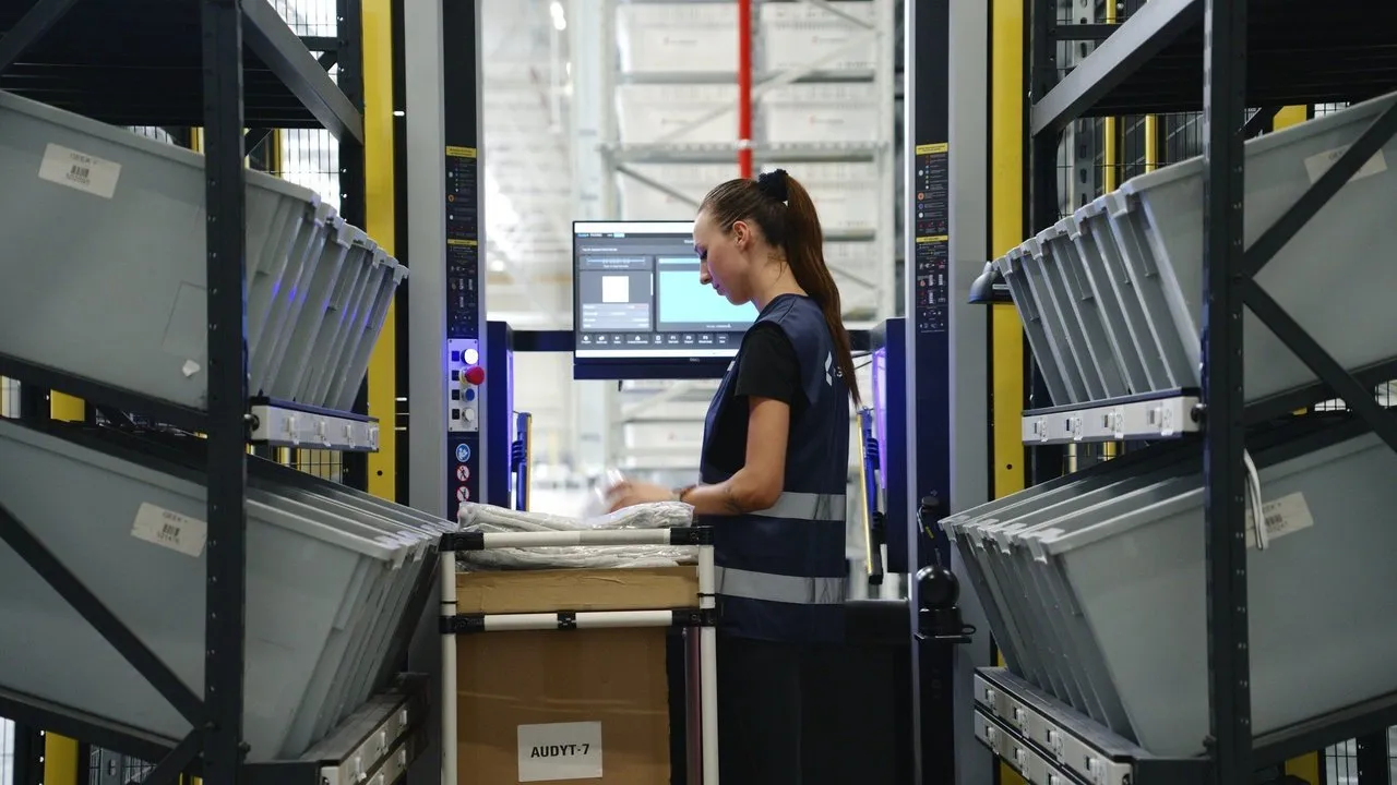 Warehouse worker using a cart beside storage racks of gray bins and a computer screen.
