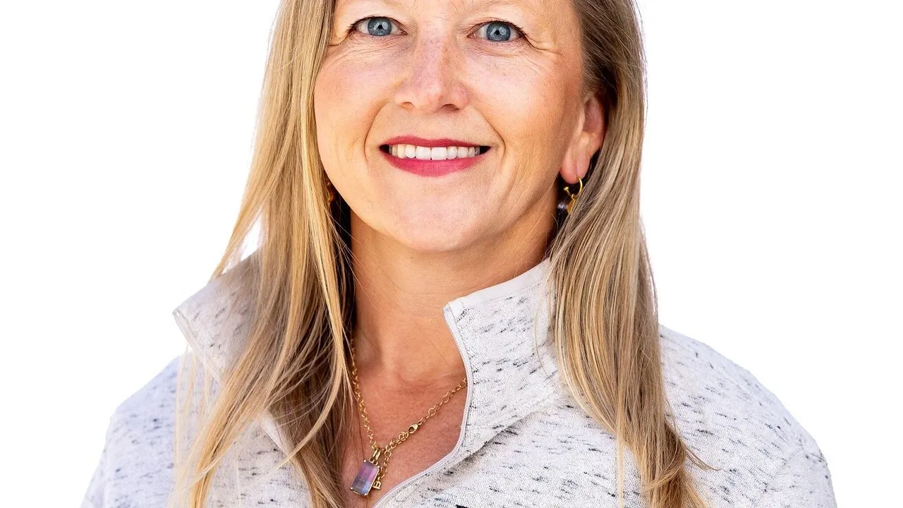 Smiling blonde woman wearing a light gray zip-up jacket and gold jewelry against a white background