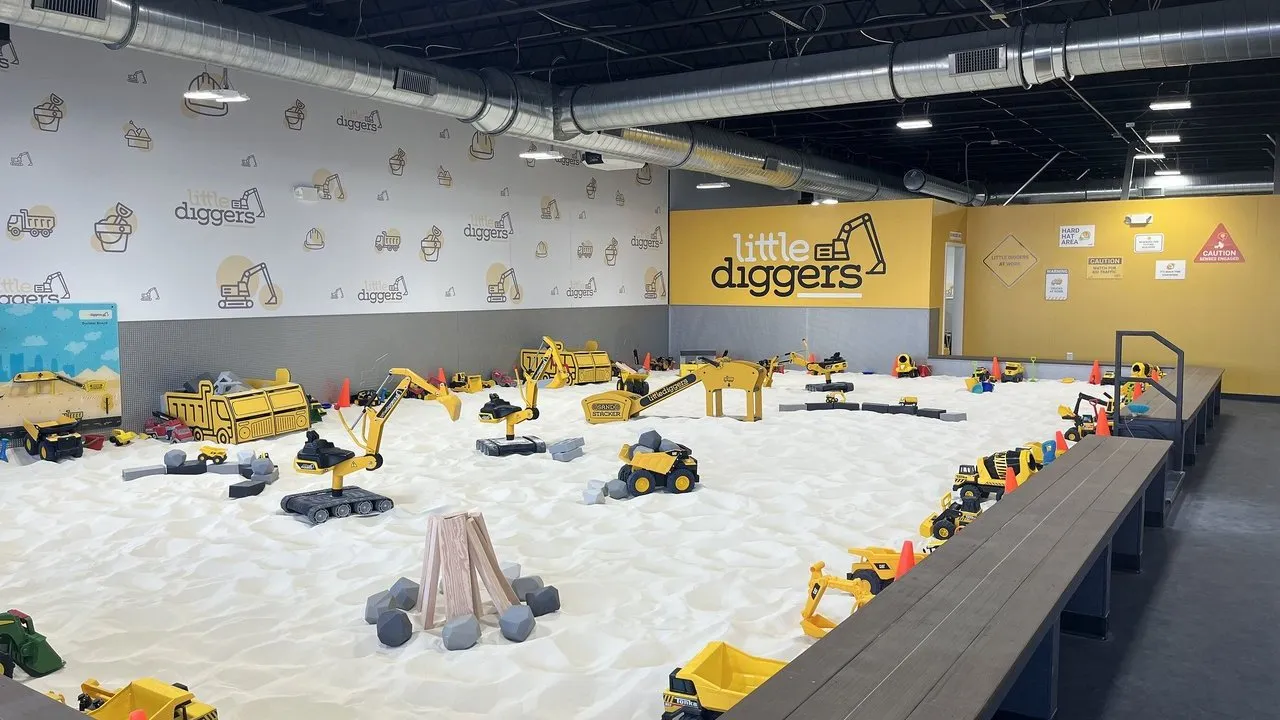 Indoor sand play area with toy construction vehicles, cones and Little Diggers signage.