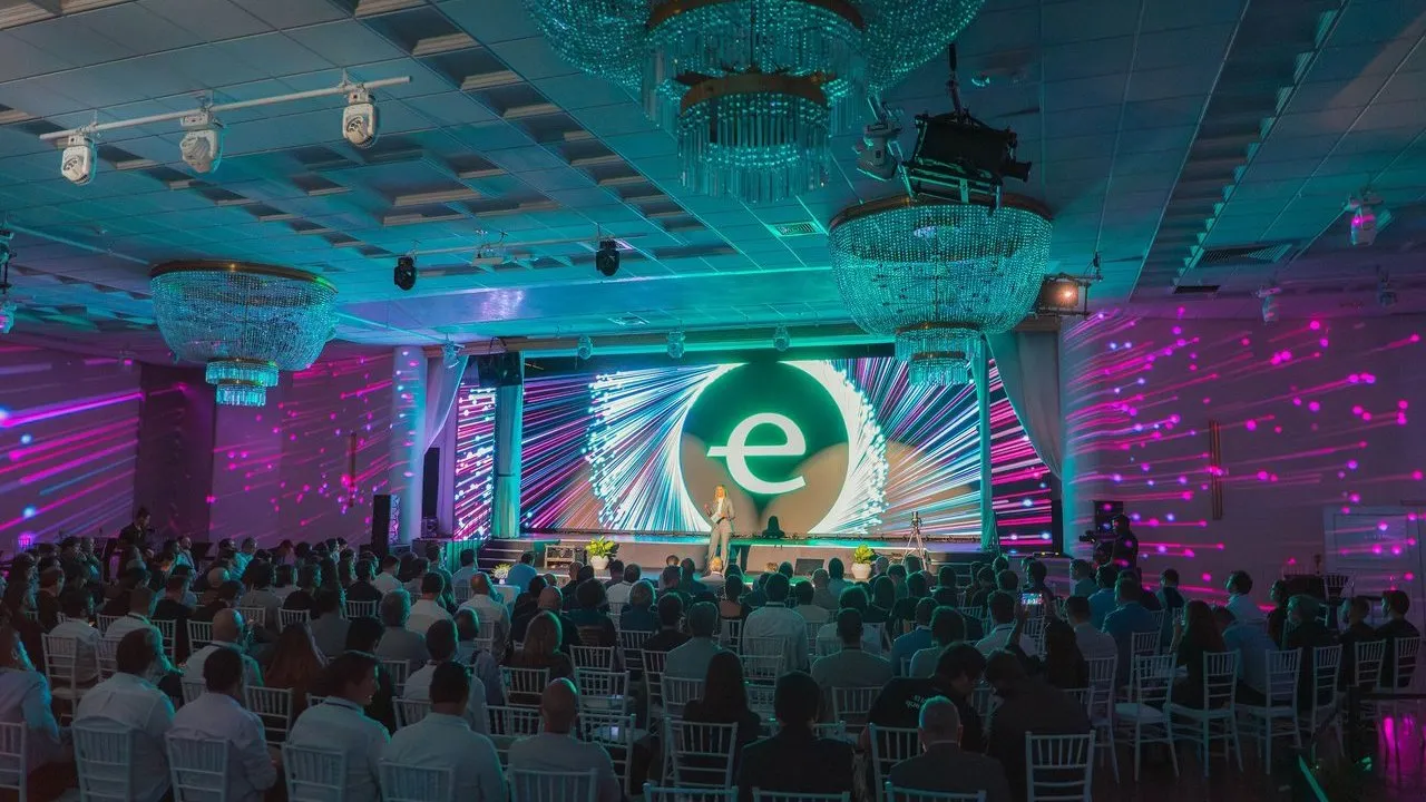 Audience seated before a brightly lit main stage with a speaker and large circular logo screen.