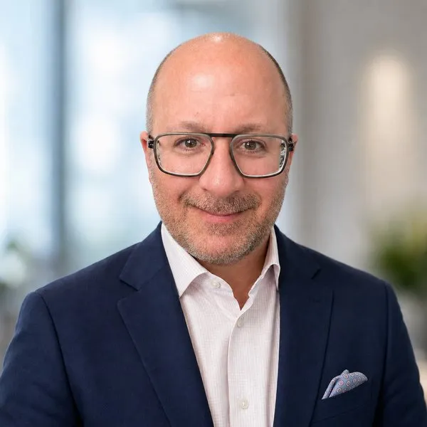 A bald man wearing glasses, a navy blazer and a white shirt smiles at the camera.