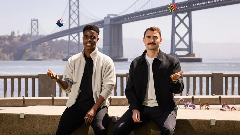 Two men juggle Rubik’s Cubes on a waterfront promenade with a suspension bridge behind them.