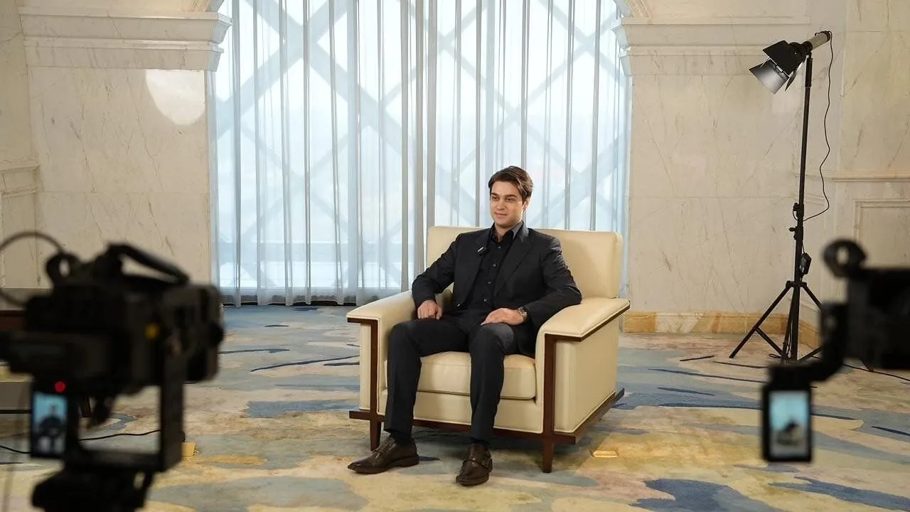 A man in a dark suit sits in an armchair between two video cameras and studio lights.