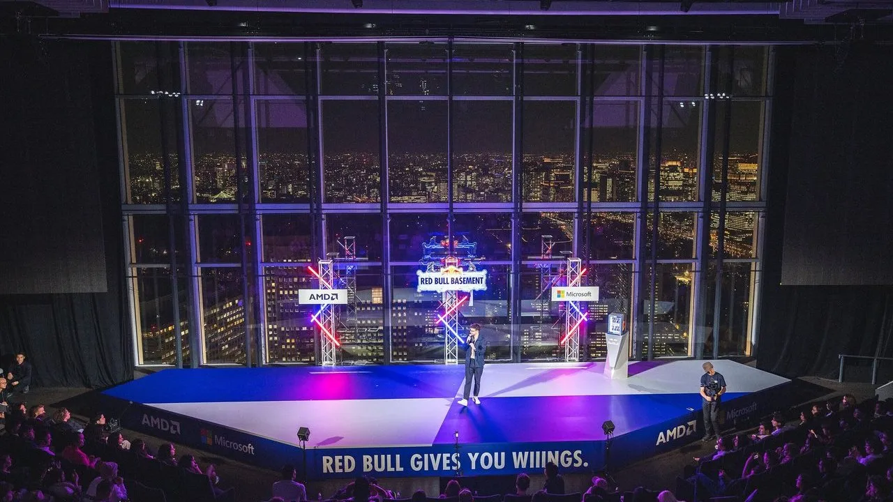Speaker on a lit Red Bull Basement stage before a nighttime city skyline and seated audience.