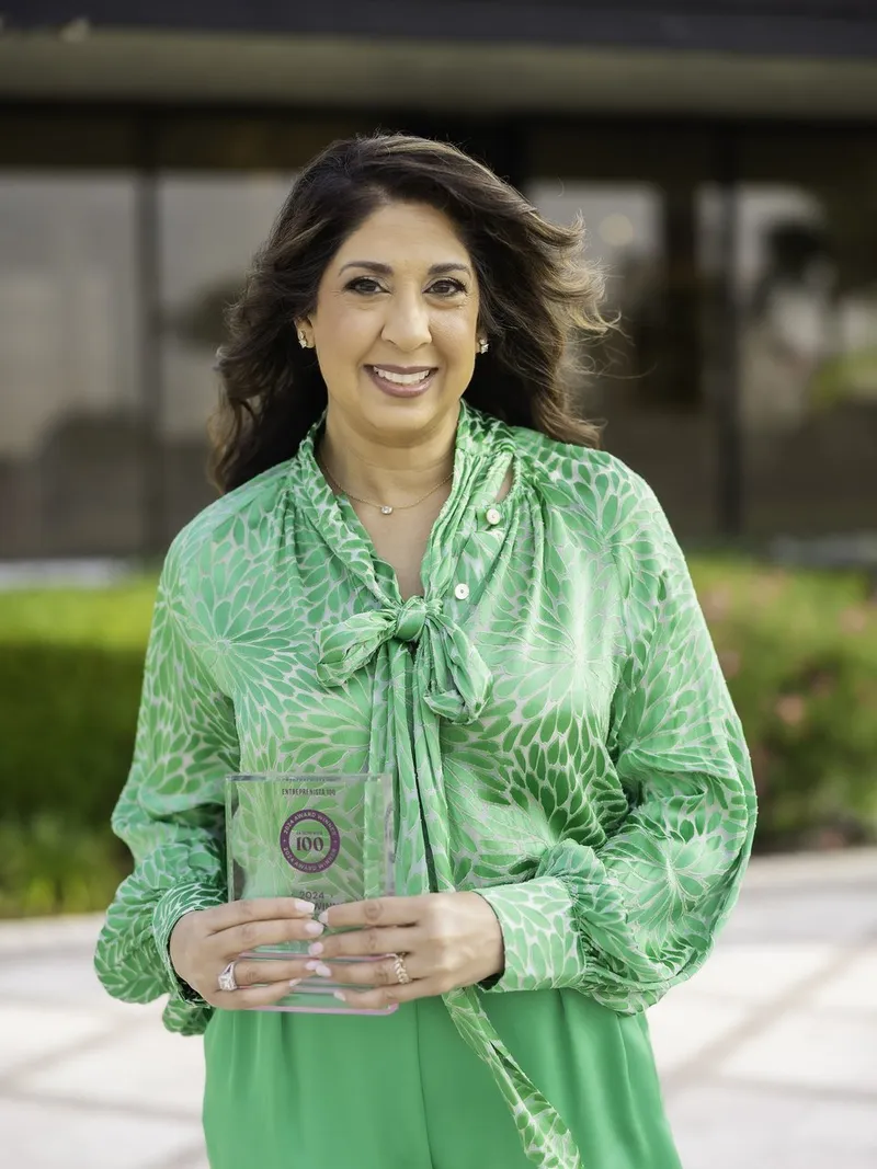 Ruchi Pinniger holds an award plaque, wearing a green patterned blouse outdoors.
