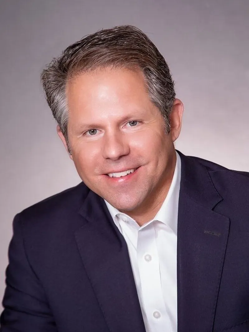 Rusty Rhodes in a navy suit jacket and white shirt against a gray background.