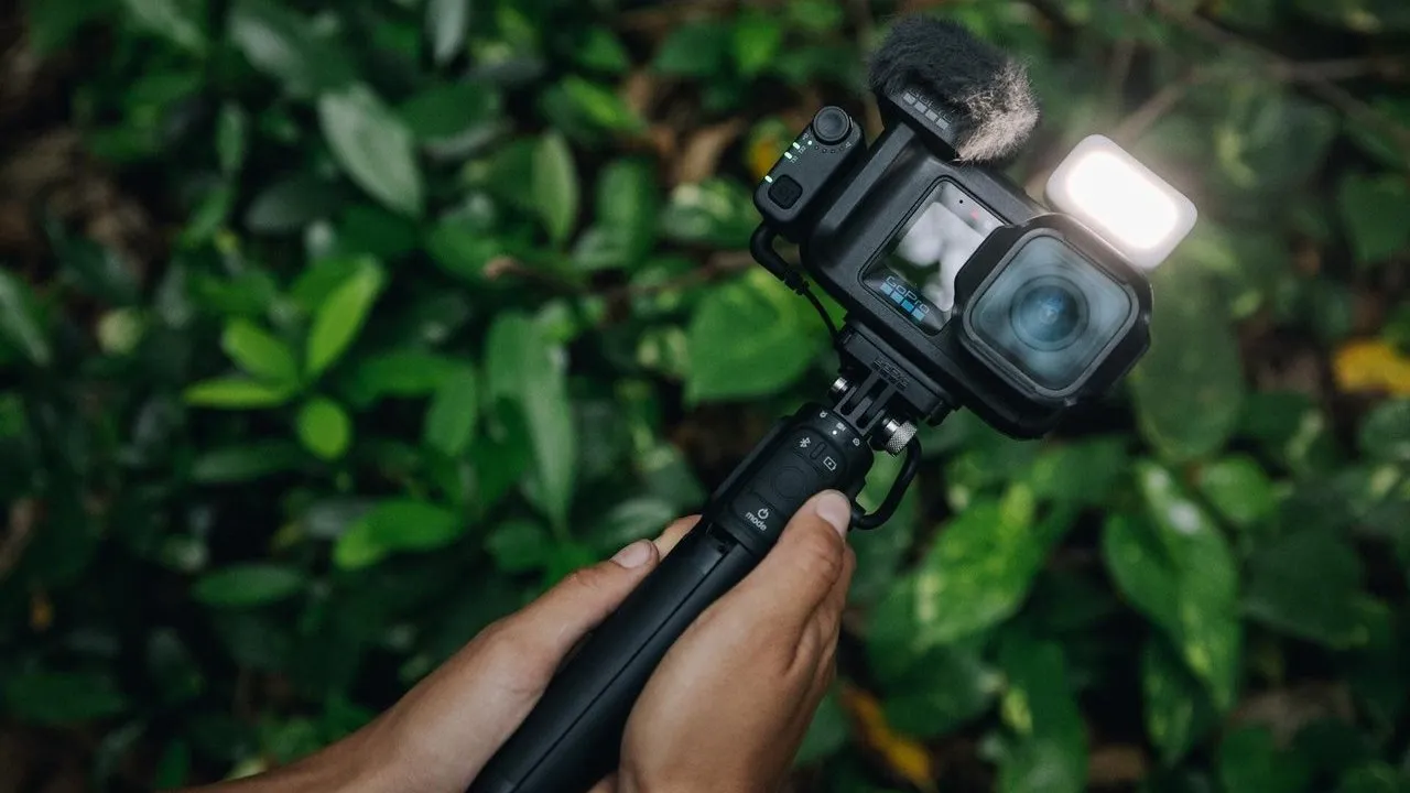Hands holding an action camera on a tripod grip with a microphone and light in a leafy forest.