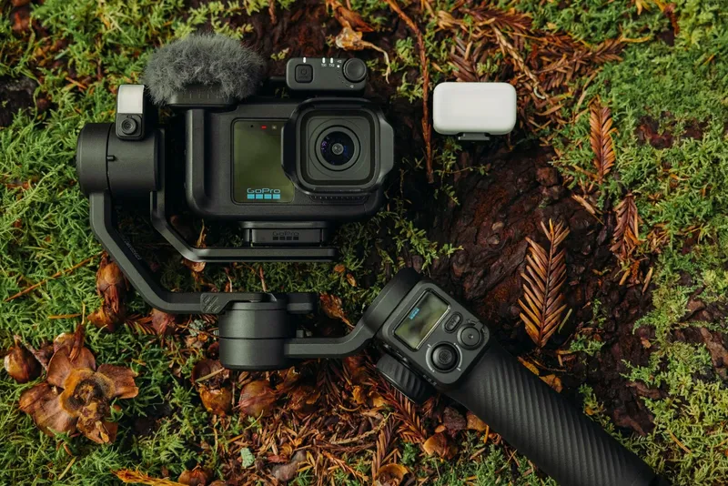 A GoPro camera mounted on a handheld gimbal beside a wireless microphone case on mossy forest ground