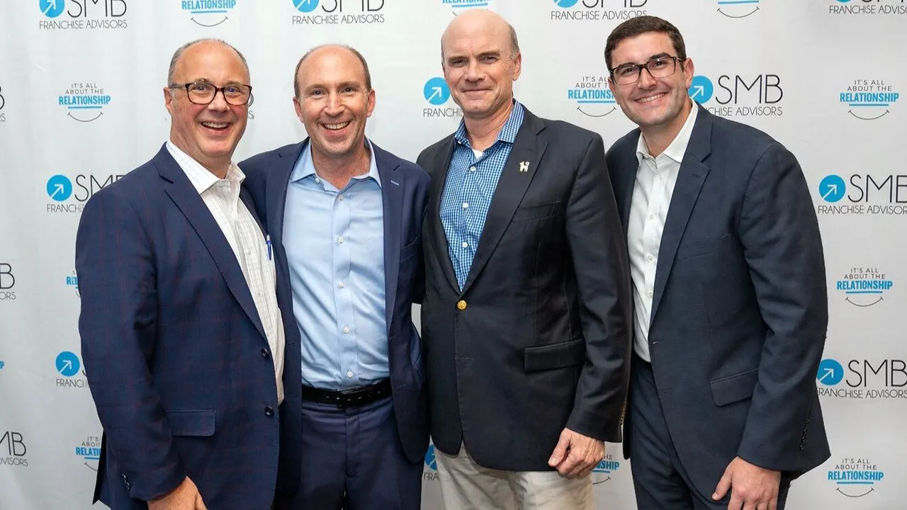 Four men in business suits pose before an SMB Franchise Advisors backdrop.