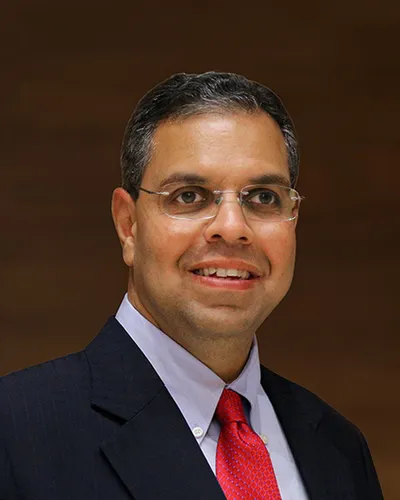Sameer Kanodia in a dark suit, glasses and red tie against a brown background.