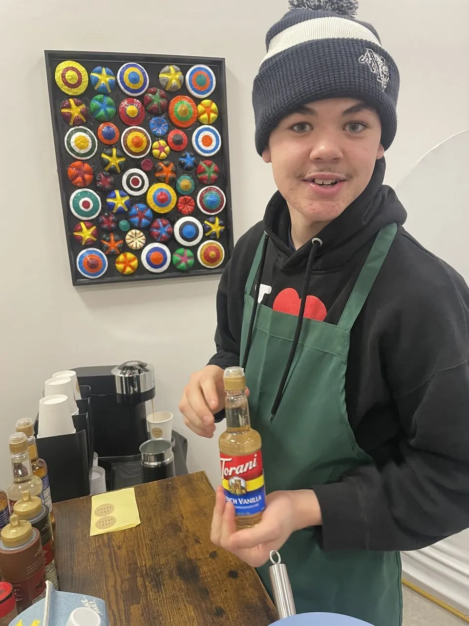 A student barista in an apron holds a vanilla syrup bottle beside coffee supplies.